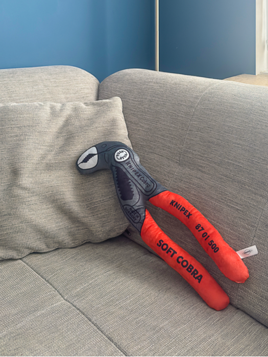 Ein Stoffspielzeug in Form einer Knipex Cobra Zange mit roten Griffen und grauem Zangenkopf, auf einem grauen Sofa liegend.