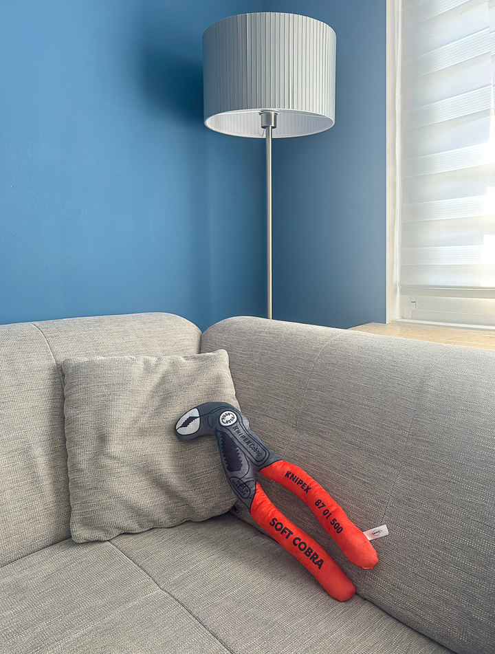 Plüsch-Version einer Knipex Cobra Wasserpumpenzange mit rotem Griff, auf einem grauen Sofa neben einem Kissen, vor einer blauen Wand