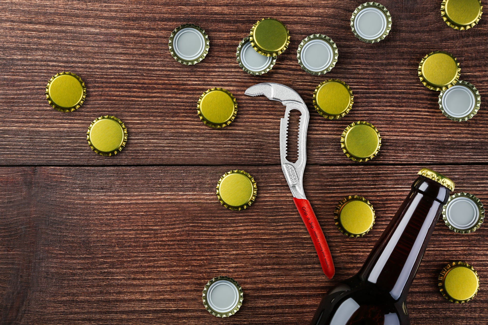 Auf einer dunklen Holzoberfläche liegen eine geöffnete Bierflasche, mehrere goldene Kronkorken und ein Flaschenöffner in Form einer kleinen Rohrzange mit rotem Griff.