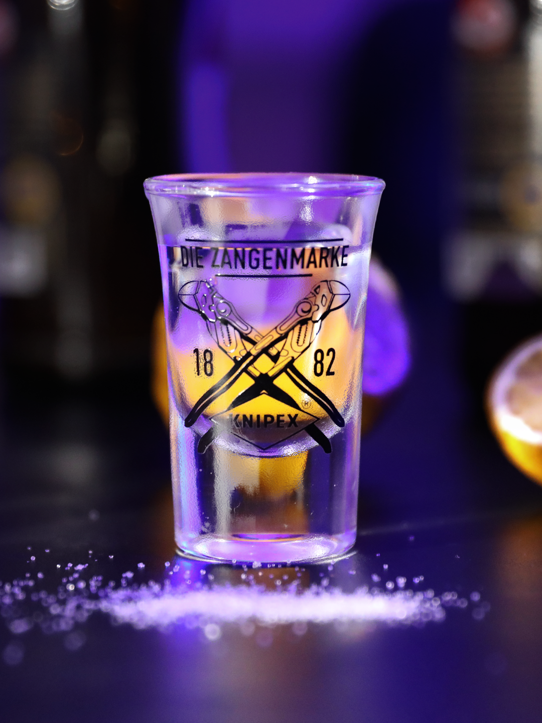 Knipex Pinnchen mit der Aufschrift "Die Zangenmarke 1882" und Knipex Logo, beleuchtet in Lila mit Zitronen im Hintergrund 