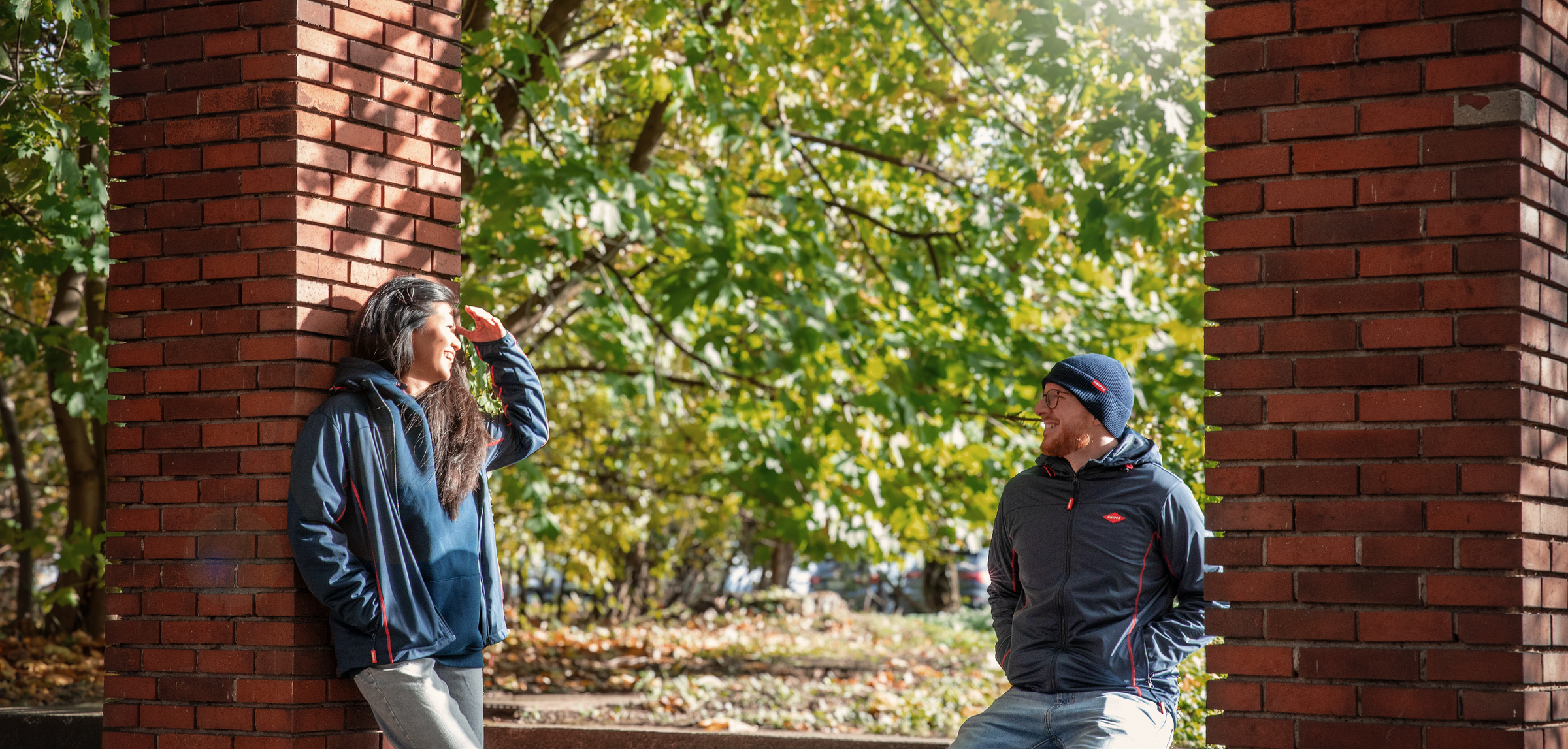 Zwei Personen tragen dunkelblaue Kleidung mit Knipex-Logo,Jacken, eine Mütze und einen Hoodie, mit Bäumen  im Hintergrund, zwischen zwei Säulen