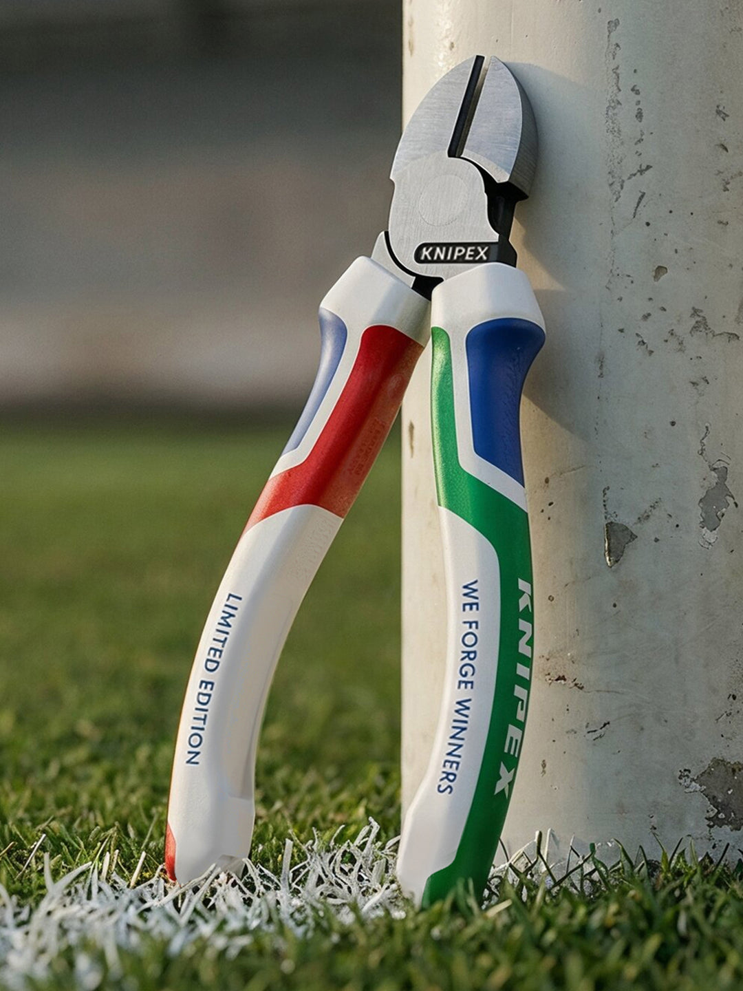 KNIPEX „WE FORGE WINNERS“ Seitenschneider in limitierter Sonderedition mit Griffen in Weiß, Blau, Grün und Rot, angelehnt an einen Pfosten auf Gras, mit präzisen, induktiv gehärteten Schneiden und ergonomischen Comfort-Griffen.