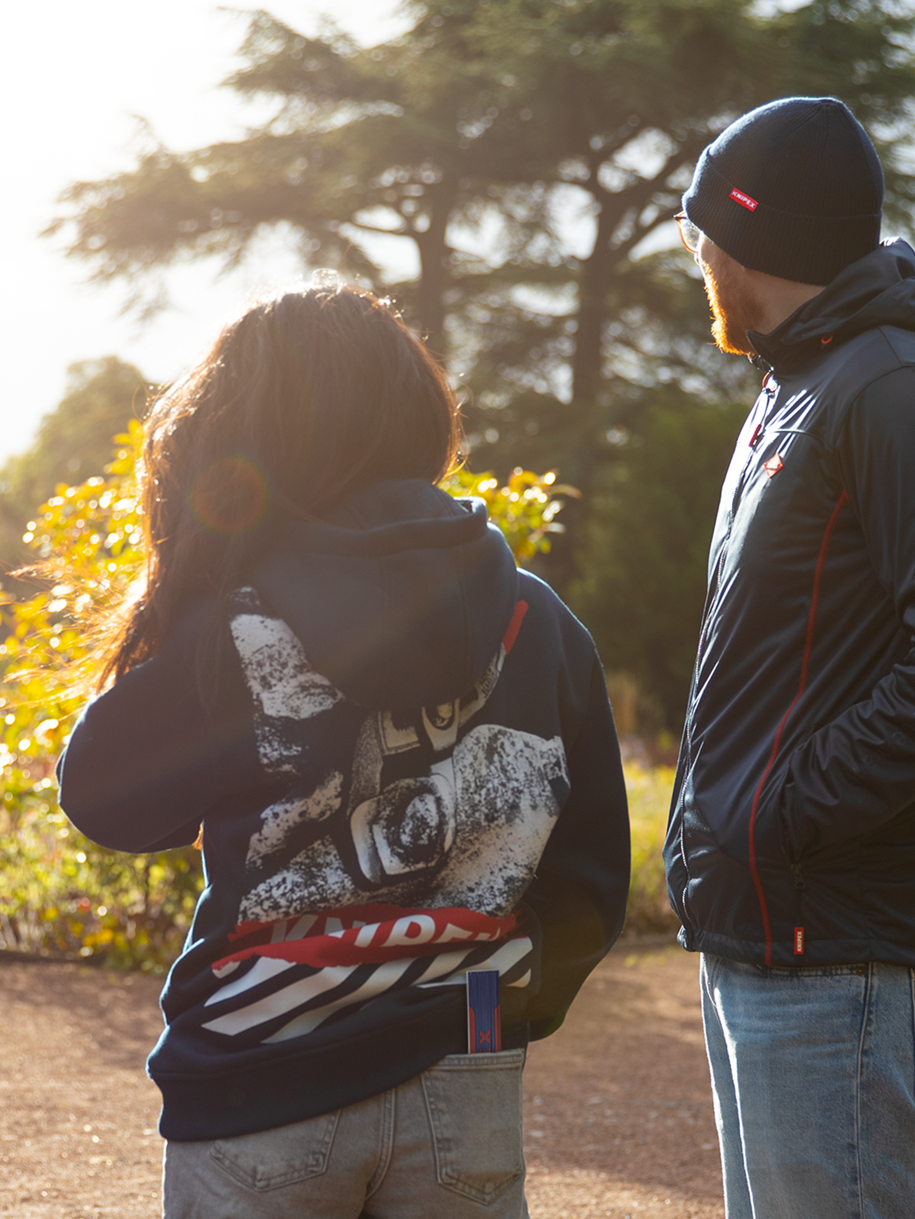 Zwei Personen tragen dunkelblaue Kleidung mit Knipex-Logo, eine Jacke, eine Mütze und einen Hoodie, mit Bäumen  im Hintergrund.