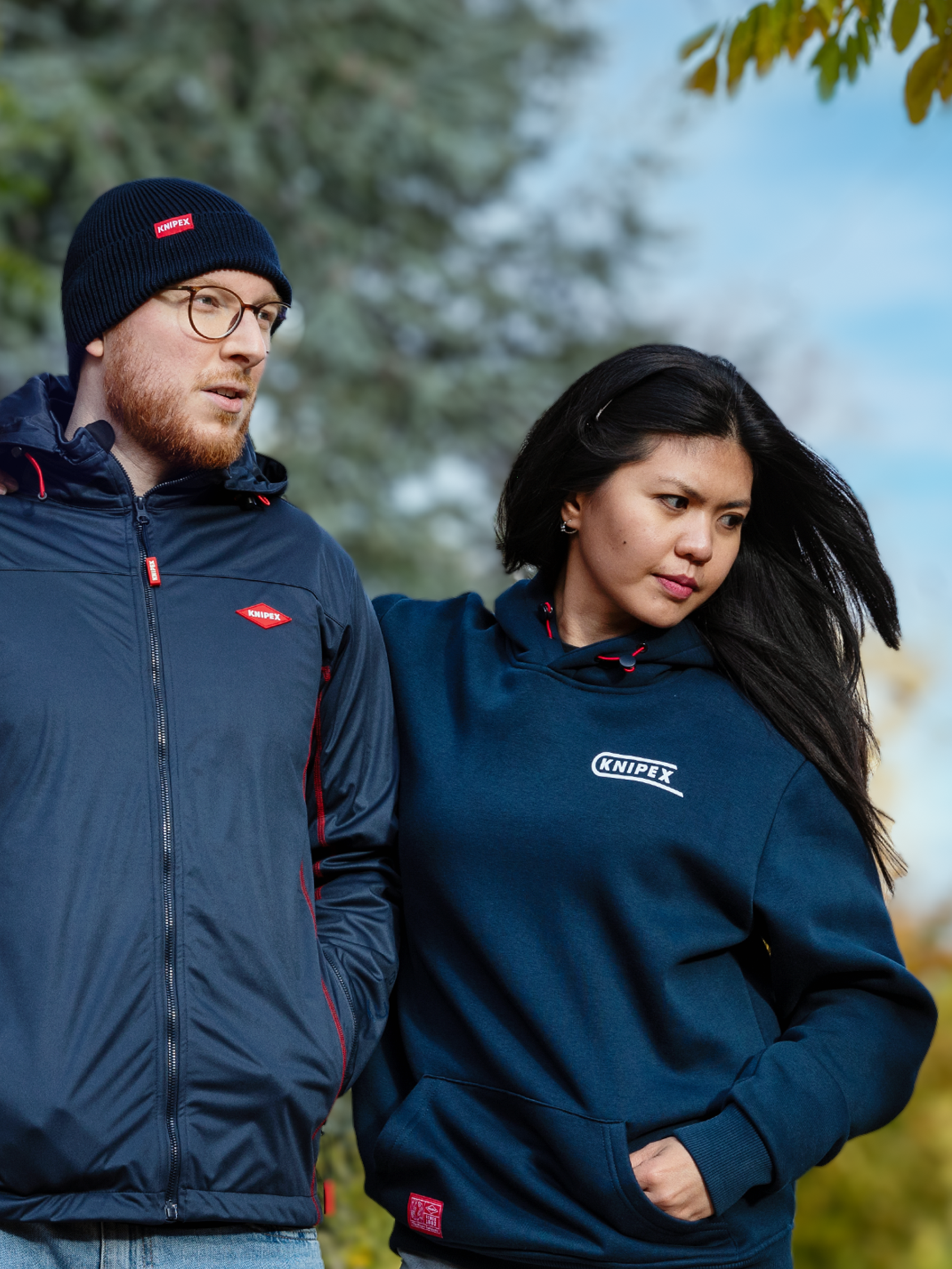 Zwei Personen in dunkelblauer Kleidung mit Knipex-Logo, eine Jacke und einem Hoodie, mit Bäumen und blauem Himmel im Hintergrund.