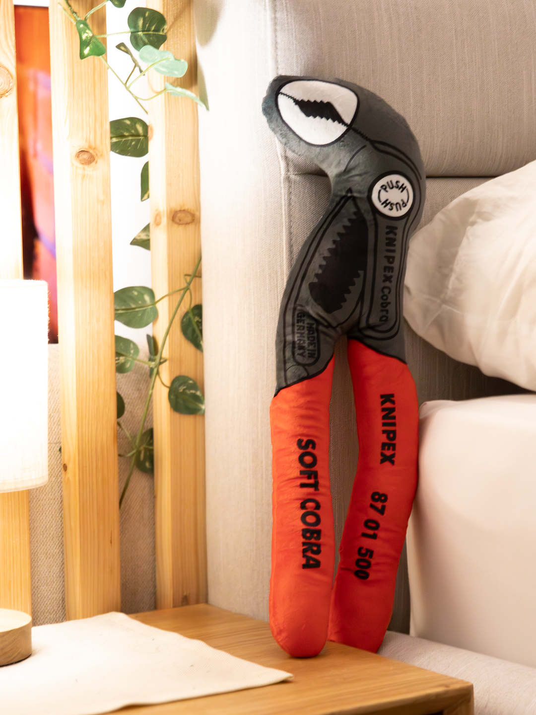 Plüsch-Version einer Knipex Cobra, rote Griffe grauer Kopf, im Schlafzimmer vor einem Bett 