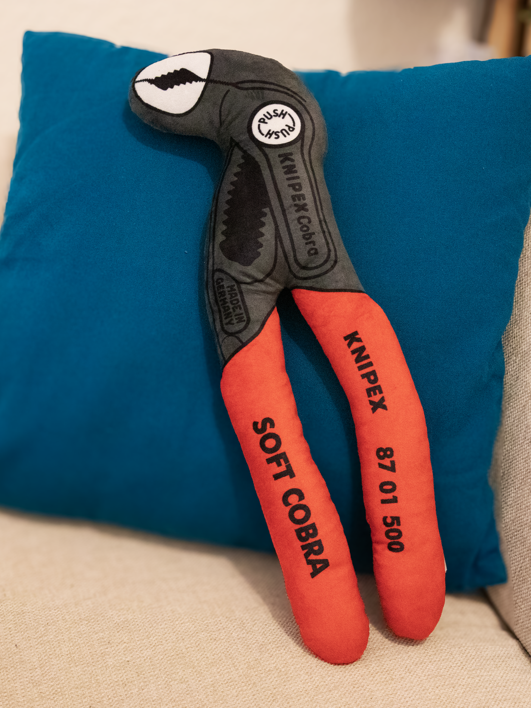 Plüsch-Version einer Knipex Cobra , rote Griffe grauer Kopf vor einem blauen Kissen  