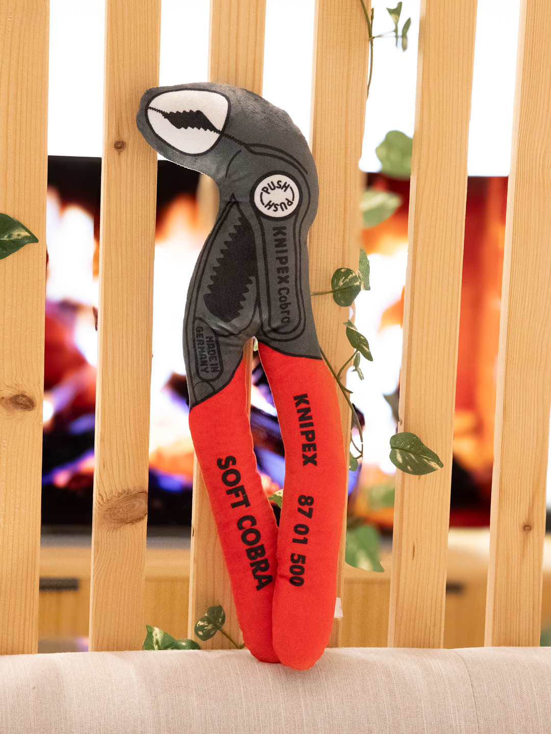 Plüsch-Version einer Knipex Cobra Wasserpumpenzange mit rotem Griff und grauem Kopf auf einem beigen Sofa vor einer Holzstrebenwand