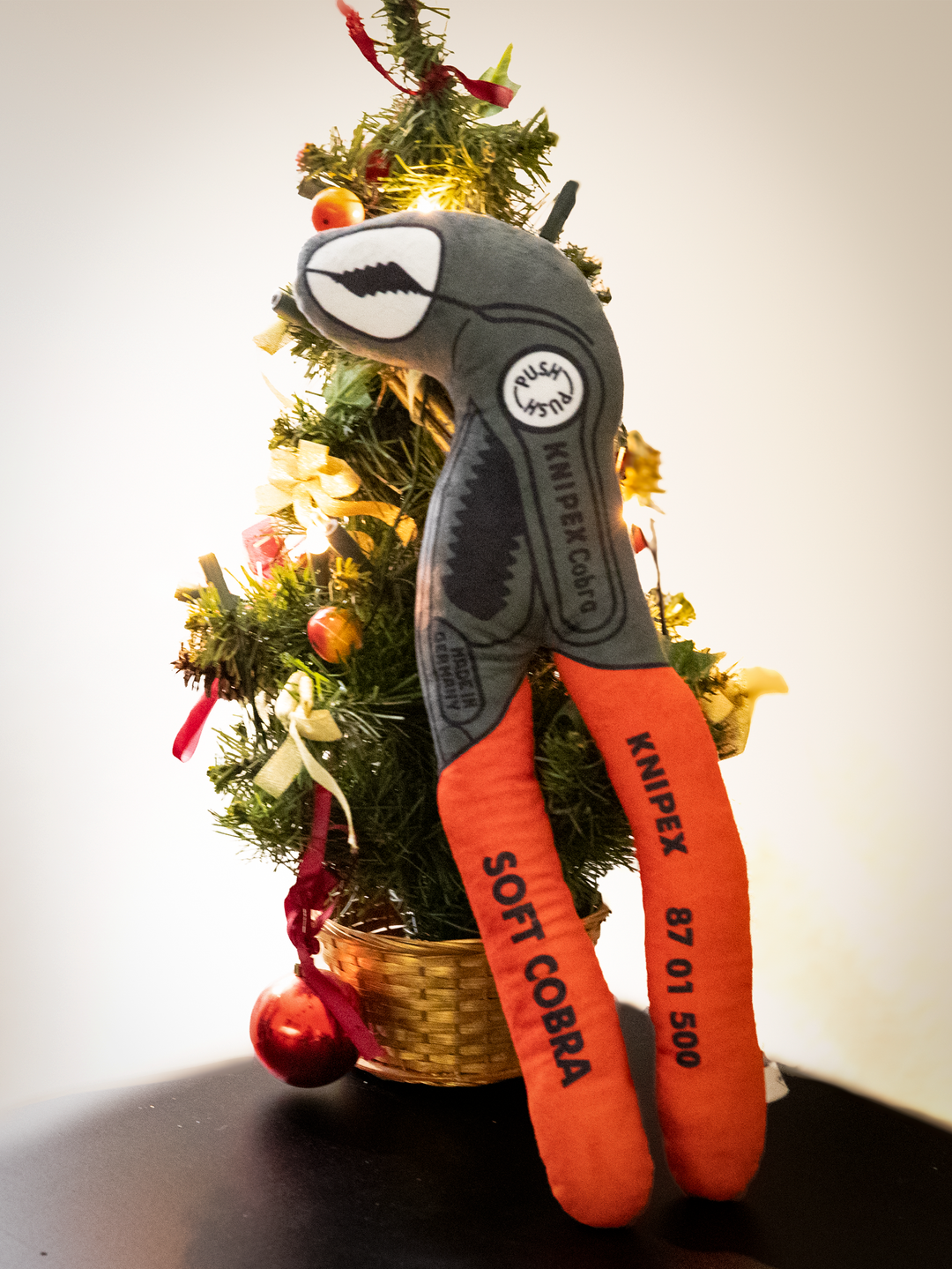 Plüsch-Version einer Knipex Cobra Wasserpumpenzange mit rotem Griff grauem Kopf, vor einem Tannenbaum
